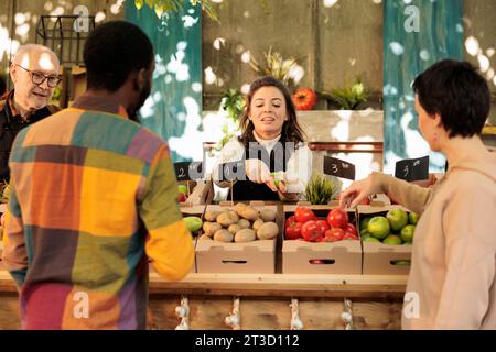 Femme souriante joyeuse offrant au client d'essayer un petit morceau de pomme tout en vendant des fruits et légumes biologiques frais au marché des agriculteurs locaux, une attention sélective. Dégustation pendant les courses Banque D'Images