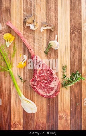 Un tak de tomahawk bien marbré sur le bloc de boucheries avec des ingrédients frais de romarin, de champignons huîtres, de fenouil et de grains de poivre. Banque D'Images