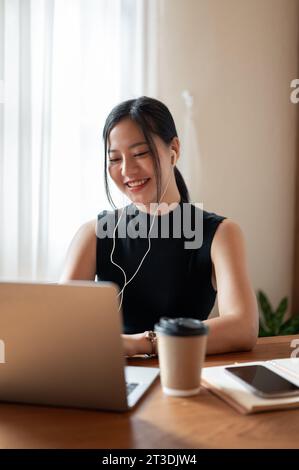 Une belle et heureuse femme asiatique millénaire portant des écouteurs et ayant une réunion en ligne via ordinateur portable avec ses collègues tout en travaillant à distance à un Banque D'Images