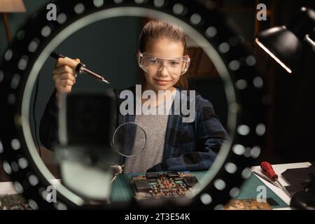 Heureuse fille souriante blogger avec lumière annulaire et smartphone streaming à la maison, parlant de carte mère à souder. Blogging, technologie et hobby Banque D'Images