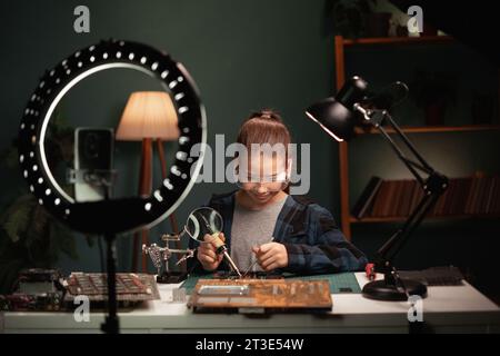 Heureuse fille souriante blogger avec lumière annulaire et smartphone streaming à la maison, parlant de carte mère à souder. Blogging, technologie et hobby Banque D'Images