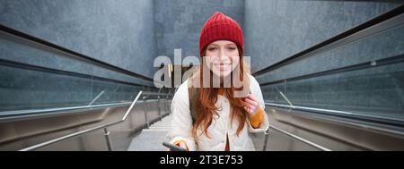 Photo en extérieur d'une jeune femme qui planifie un itinéraire, suit la carte sur l'application pour smartphone, monte les escaliers avec un sac à dos et sourit Banque D'Images