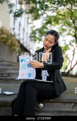 Une femme d'affaires asiatique millénaire occupée, stressée et insatisfaite vérifie les documents et se plaint du problème au téléphone tout en étant assise Banque D'Images