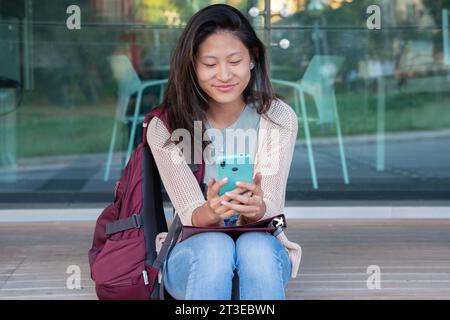 jeune femme asiatique assise naviguant sur internet sur smartphone à l'extérieur. Banque D'Images