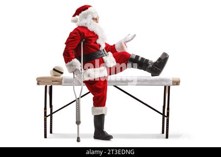 Père noël avec une orthèse de pied et béquille assis sur un lit médical et gestuant avec la main isolé sur fond blanc Banque D'Images