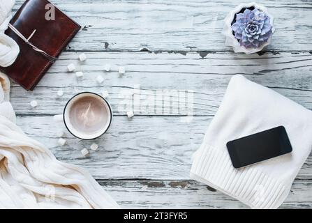 Au-dessus de l'automne ou de l'hiver arrière-plan photographié de dessus avec du cacao chaud, plante d'intérieur, téléphone portable, livre, et jeter couverture sur le bois blanc rustique dos Banque D'Images