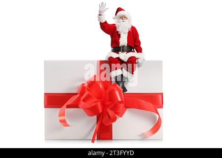 Portrait en longueur du père noël assis sur un grand cadeau et agitant isolé sur fond blanc Banque D'Images