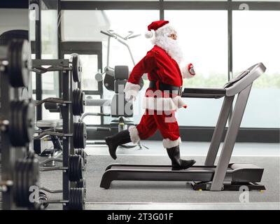 Photo de profil pleine longueur du Père Noël courant sur un tapis roulant dans une salle de gym Banque D'Images