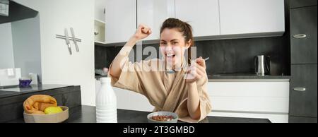 Portrait de jeune femme heureuse et rieuse mangeant des céréales avec du lait, triomphant, prenant le petit déjeuner et se sentant excitée, concept du matin énergique Banque D'Images