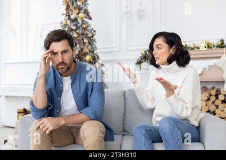 Conflit d'un jeune couple, famille sur les vacances du nouvel an. Un jeune homme et une femme sont assis sur le canapé et se disputent. La femme crie et accuse le mari, le mari tient sa tête. Banque D'Images