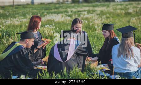 Diplômés en costumes noirs mangeant de la pizza dans une prairie de la ville. Banque D'Images