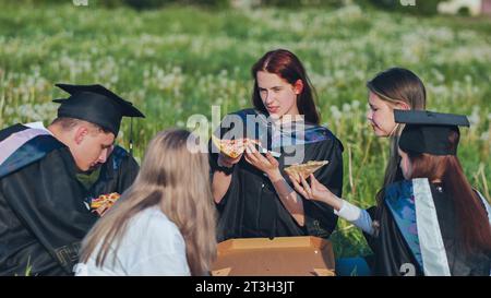 Diplômés en costumes noirs mangeant de la pizza dans une prairie de la ville. Banque D'Images
