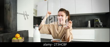 Portrait de jeune femme heureuse et rieuse mangeant des céréales avec du lait, triomphant, prenant le petit déjeuner et se sentant excitée, concept du matin énergique Banque D'Images