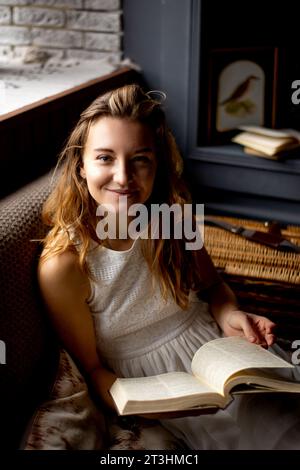 Assise sur le canapé, une jeune femme à l'apparence attrayante et aux cheveux longs sourit chaleureusement tout en tenant un livre. La photo parle du plaisir Banque D'Images