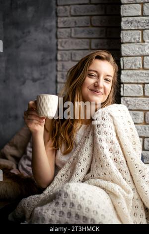 Une jeune femme joyeuse tient une tasse à café, son sourire reflétant la chaleur de sa boisson. Cette image incarne le confort des rituels matinaux et du JO Banque D'Images