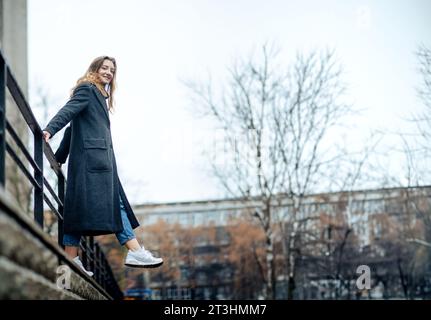 Une jeune femme dans un long manteau gris près des écoutes blanches fait un pas dans l'abîme. Elle regarde la caméra et se trouve dans une rue de la ville Banque D'Images