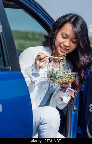 Une jeune belle coréenne en vêtements blancs mange une salade fraîche dans la voiture. Une femme asiatique attrayante a une collation tout en voyageant. Sain et Banque D'Images