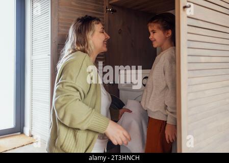 Une maman attrayante et son bébé mignon font les tâches ménagères. Une jeune femme et sa fille chargent une machine à laver. Une adorable mère l'embrasse éclairée Banque D'Images