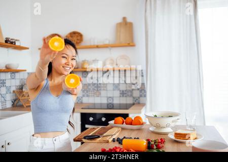 Une jeune femme asiatique séduisante sourit et montre des oranges à la caméra. Une charmante dame coréenne prépare du jus d'orange. Une belle fille thaïlandaise se prépare Banque D'Images