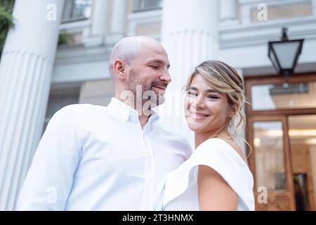 Marié attrayant et charmante mariée sur fond de bâtiment avec de grandes colonnes. Jeune homme en chemise blanche embrasse la belle femme en élégant dr Banque D'Images