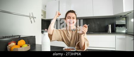 Portrait d'une jeune femme enthousiaste mangeant des céréales avec du lait, paraissant excitée et heureuse, assise près du plan de travail de la cuisine et prenant le petit déjeuner, élevant Banque D'Images