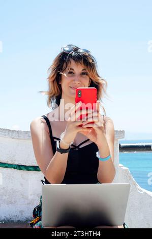 femme assise regardant la caméra tout en tapant avec le téléphone portable Banque D'Images