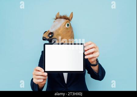 Un jeune homme joyeux avec masque de cheval en costume isolé sur fond bleu démontrant l'écran blanc de sa tablette Banque D'Images