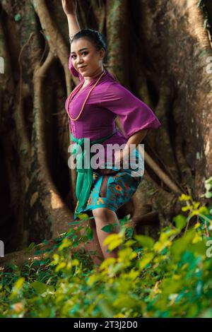 Femme asiatique prend une pose de danse dans la jungle tout en portant une robe violette et un collier d'or avec un beau sourire sur son visage Banque D'Images