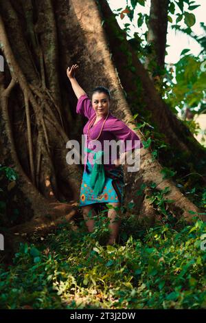 Une femme indonésienne pose courageusement en levant les mains dans une robe violette tout en se tenant debout dans la forêt Banque D'Images