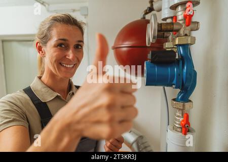 Femme ingénieur en chauffage montre le pouce vers le haut dans une chaufferie avec un vieux système de chauffage au gaz avec liste de contrôle sur un presse-papiers. Obligation de remplacement du réchauffeur à gaz Banque D'Images