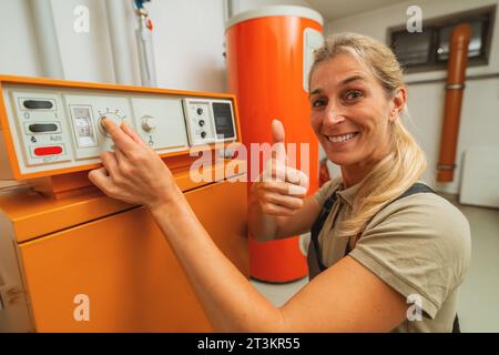 Heureuse femme ingénieur en chauffage montre le pouce vers le haut dans une chaufferie avec un vieux système de chauffage au gaz avec liste de contrôle sur un presse-papiers. Obli de remplacement du réchauffeur à gaz Banque D'Images