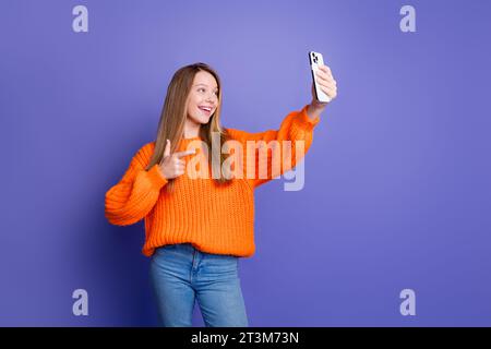 Jeune adolescente gestuelle doigt symbole abonnés photo sont cool prend selfie shot à l'aide d'un smartphone isolé sur fond de couleur violette Banque D'Images