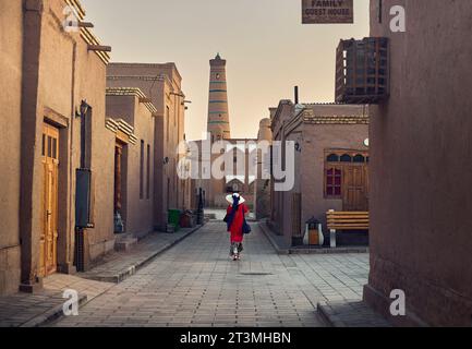 Femme en robe rouge marchant dans la vieille ville itchan kala à Khiva au lever du soleil contre Islam Khoja Minaret en Ouzbékistan. Banque D'Images