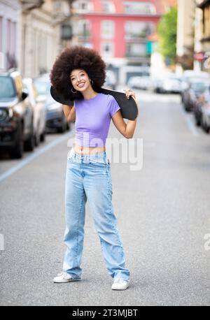 Corps entier de jeune femme afro-américaine positive avec les cheveux bouclés posant avec skateboard sur la rue de la ville Banque D'Images