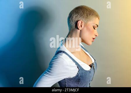 Vue de côté de jeune modèle féminin assuré avec les cheveux blonds courts et le maquillage dans les vêtements décontractés regardant loin sur fond dégradé Banque D'Images