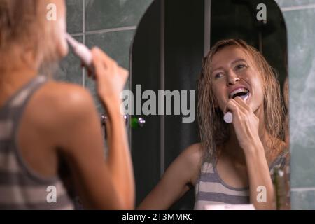 Jeune femme blonde se brosse les dents avec une brosse à dents électrique blanche dans la salle de bain regardant dans le miroir. L'hygiène, la notion de soins corporels. Banque D'Images