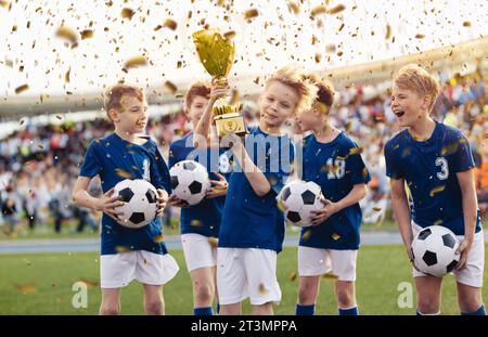 Groupe d'écoliers célébrant ensemble la victoire d'une compétition sur un terrain de football en confettis. L'équipe de football se lève le trophée de coupe d'or à l'arrière Banque D'Images