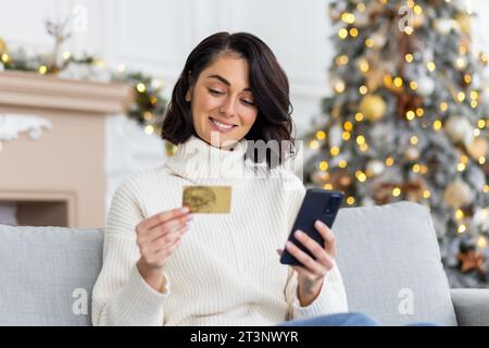 Une jeune belle femme est assise sur le canapé à la maison, tenant une carte de crédit et le téléphone et faisant des achats en ligne à la veille du nouvel an et des vacances de Noël. Banque D'Images