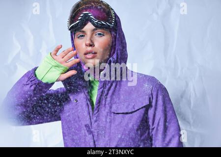 jolie jeune femme en balaclava, veste d'hiver violette et googles de ski posant le jour gris et enneigé Banque D'Images