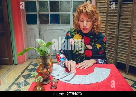 Vue de face de l'élégante femme de lecteur de tarot latin blond-poil assise à la maison prenant la carte du pont de tarot, commençant la consultation avec l'univers, copie sp Banque D'Images