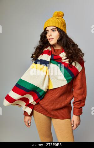 femme bouclée dans le chapeau de bobble tricoté et pull avec écharpe dépouillée sur le dessus posant sur fond gris Banque D'Images