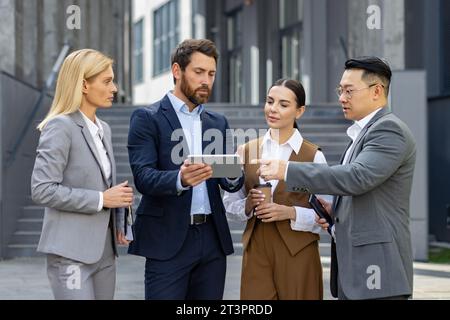 Quatre gens d'affaires discutant des résultats financiers de la réalisation, des hommes et des femmes mûrs à l'extérieur de l'immeuble de bureaux discutant de la stratégie de succès, équipe diversifiée d'hommes d'affaires et de femmes d'affaires en costumes Banque D'Images