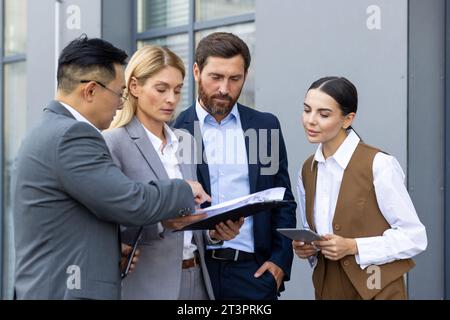 Quatre gens d'affaires discutant des résultats financiers de la réalisation, des hommes et des femmes mûrs à l'extérieur de l'immeuble de bureaux discutant de la stratégie de succès, équipe diversifiée d'hommes d'affaires et de femmes d'affaires en costumes Banque D'Images
