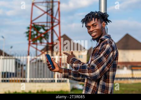 Beau gars noir avec des dreadlocks, portant un t-shirt tenant et pointant vers un smartphone. Homme africain utilisant la technologie sans fil Banque D'Images