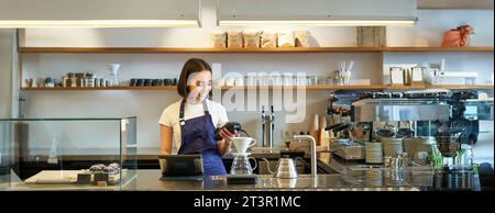 Portrait d'une jeune fille étudiante travaille comme barista, tient la machine à cartes de crédit POS, debout au comptoir avec le terminal et le kit de préparation Banque D'Images