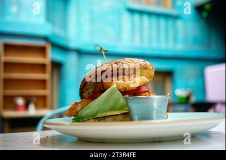 Hamburger de poulet empilé, apprécié dans un restaurant chic niché dans une cour ouverte aux murs bleus vifs. Banque D'Images