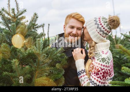 Couple choisissant arbre de Noël ensemble à un marché, elle murmure quelque chose à son oreille Banque D'Images