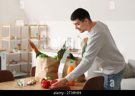 Jeune homme prenant des légumes frais de sacs d'épicerie à table dans la cuisine Banque D'Images