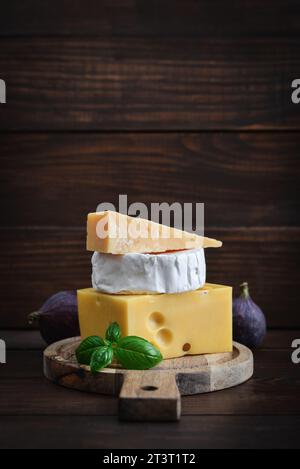Différents types de fromage : camembert, Maasdam, parmesan - sur une planche à découper avec du basilic frais et des figues sur un fond en bois Banque D'Images