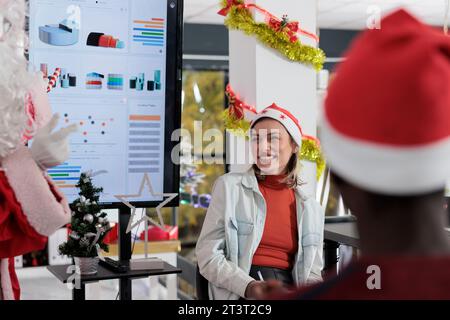 Femme d'affaires s'amusant pendant la réunion d'affaires dans la salle de conseil décorée de Noël tout en prêtant attention à l'orateur public habillé comme le Père Noël. Employé riant, appréciant la conférence de l'entreprise Banque D'Images